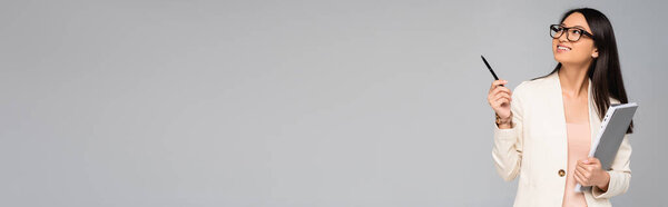 horizontal image of asian businesswoman in eyeglasses looking away and pointing with pen isolated on grey