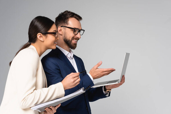young businessman pointing with hand at laptop near asian colleague holding folder isolated on grey