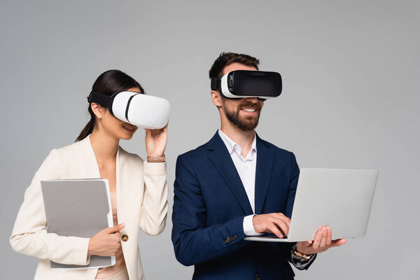 businesswoman touching vr headset near businessman using laptop isolated on grey
