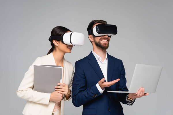 businessman pointing at laptop while using vr headsets together with business partner isolated on grey