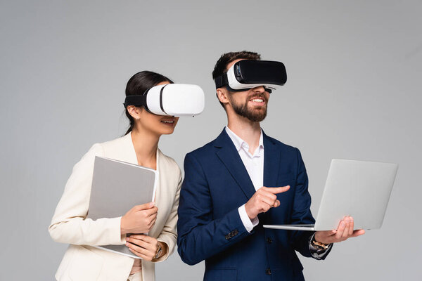 businesswoman holding folder while using vr headsets together with colleague pointing with finger at laptop isolated on grey