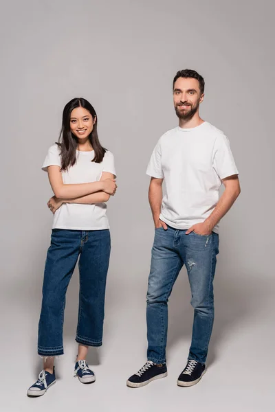 穿着白色T恤衫 牛仔裤和胶鞋的时髦的跨种族夫妇 手插在口袋里 双臂交叉 呈灰色 — 图库照片