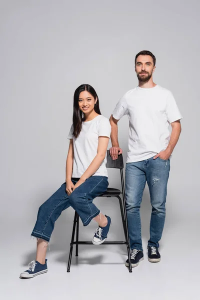 穿着白色T恤和牛仔裤的黑发亚洲女人坐在靠近时髦男友的椅子上 手挽手站在灰色口袋里 — 图库照片