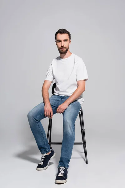 Barbudo Jovem Jeans Camiseta Branca Olhando Para Câmera Enquanto Sentado — Fotografia de Stock