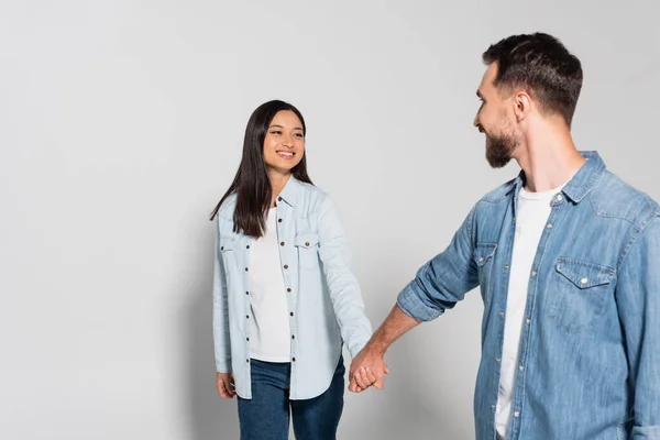 young interracial couple in denim shirts holding hands while looking at each other on grey