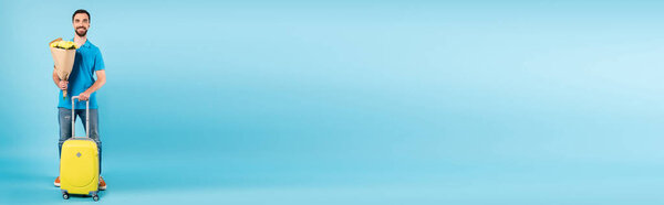 горизонтальная концепция туриста, стоящего с желтым чемоданом и держащего букет на синем