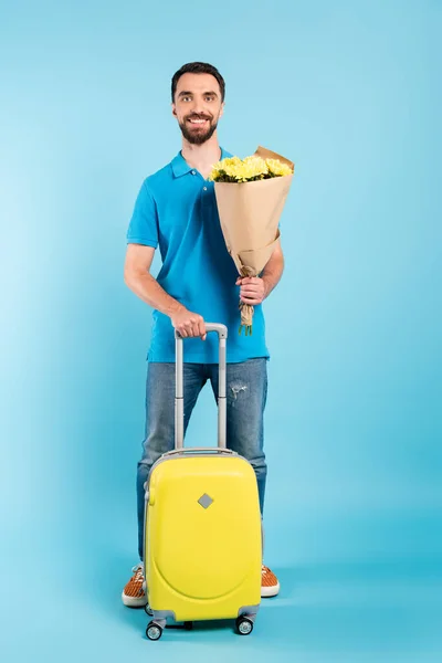 带着黄色行李箱的留胡子游客一边拿着花束一边看着蓝色的相机 — 图库照片
