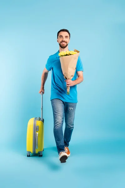 穿着马球T恤和牛仔裤的年轻游客 提着黄色的花束走在蓝色的行李箱上 — 图库照片
