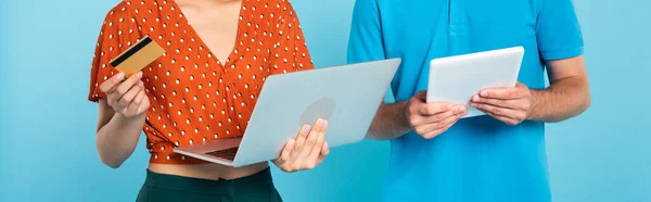 cropped view of woman in red blouse holding credit card and laptop near man with digital tablet on blue, panoramic concept