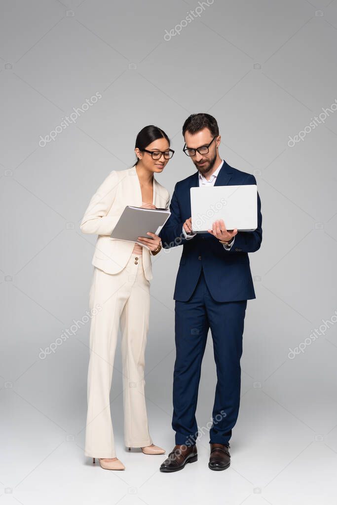 full length view of interracial business colleagues with laptop and folder on grey 