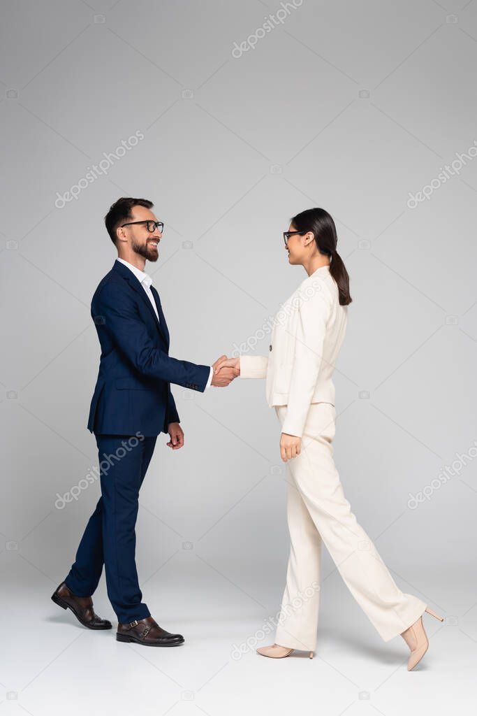 full length view of interracial business colleagues in formal wear shaking hands on grey