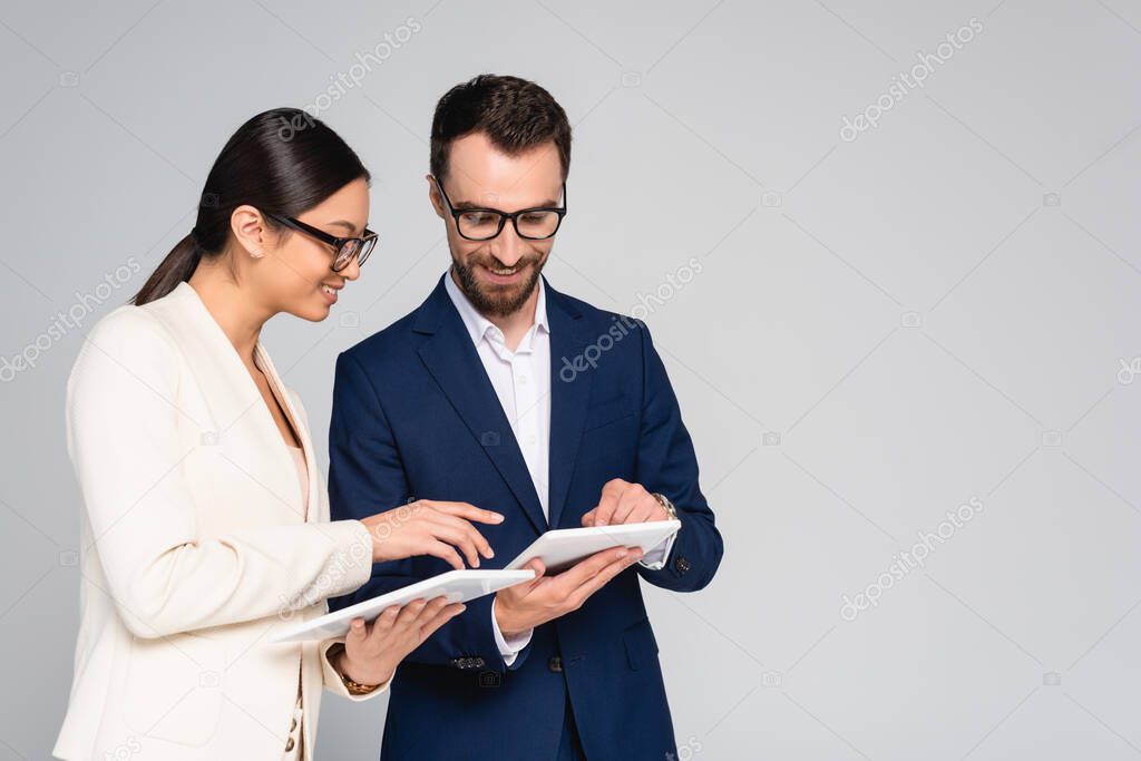 interracial couple of business colleagues in blazers and eyeglasses using digital tablets isolated on grey