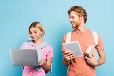 redhead student holding books and digital tablet while looking at blonde friend using laptop on blue clipart