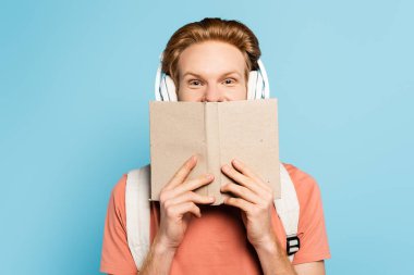 Kablosuz kulaklıklı kızıl saçlı öğrenci yüzünü mavi kitapla kapatıyor. 