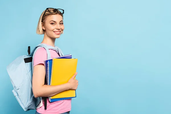 young blonde student with backpack holding notebooks on blue