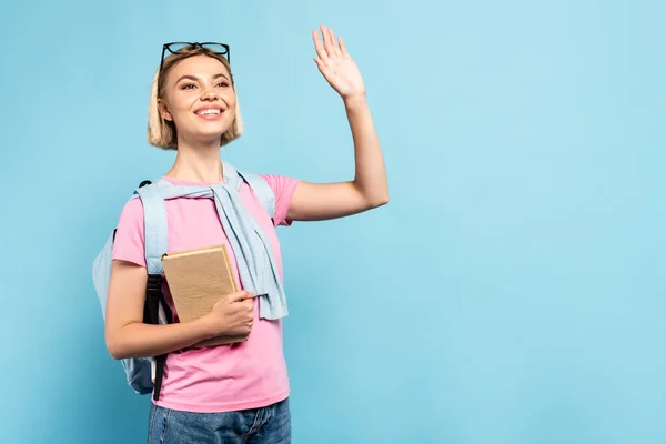 Young Blonde Student Backpack Holding Book Waving Hand Blue — Stock Photo, Image