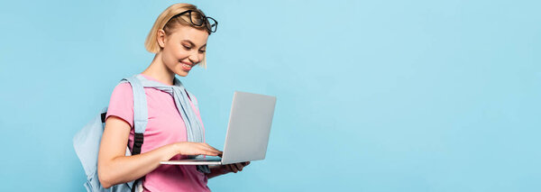 panoramic concept of young blonde student with backpack using laptop isolated on blue