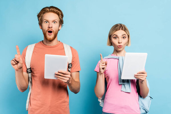 удивленные молодые студенты, держащие цифровые таблетки и показывающие знак идеи на синем