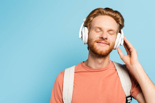 bearded and redhead student with closed eyes touching wireless headphones on blue 