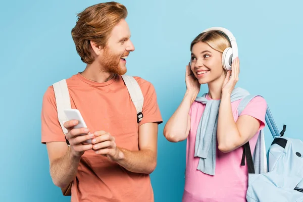 金髪の女がワイヤレスヘッドフォンで音楽を聴きながら青でスマートフォンで学生を見る — ストック写真