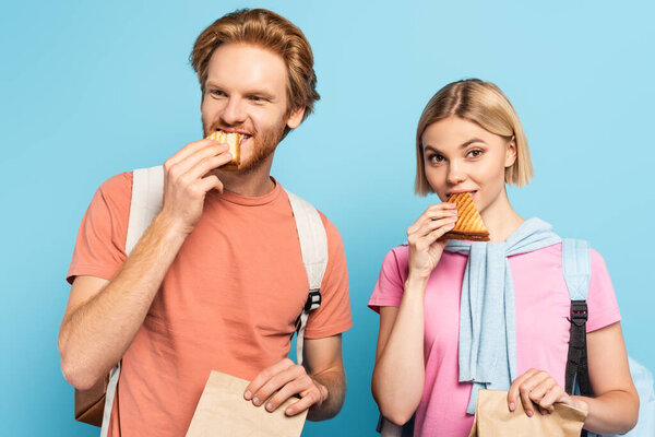 молодые студенты держат бумажные пакеты и едят сэндвичи на голубом