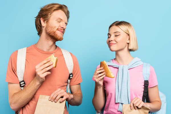 Молодые студенты с закрытыми глазами держат бумажные пакеты и сэндвичи на голубом