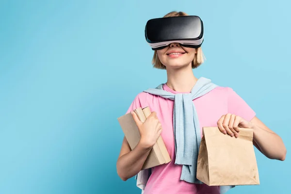 young blonde student in virtual reality headset holding books and paper bag on blue