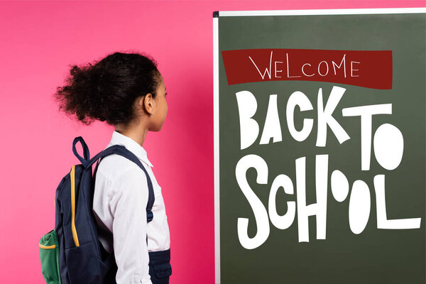 african american schoolkid looking at chalkboard with welcome back to school lettering on pink