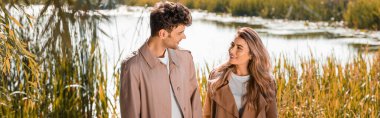 panoramic shot of pleased woman and man in trench coats looking at each other near lake in autumnal park clipart