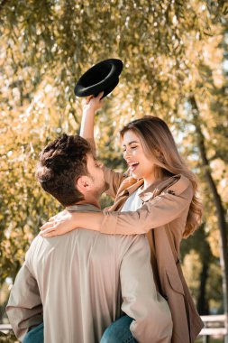 Sonbahar parkında şapkalı heyecanlı bir kadını kollarında tutan adam.
