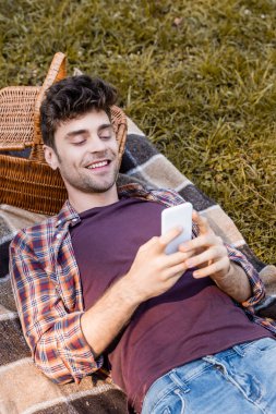 Adam hasır sepetin yanında battaniyenin üzerinde yatarken akıllı telefon kullanıyor. 