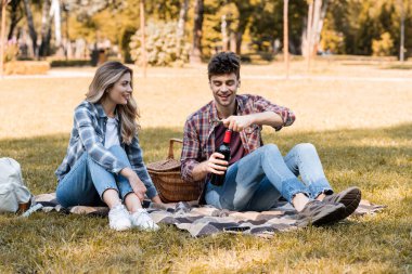 Kadının yanında elinde kırmızı şarap şişesi olan bir adam parkta battaniyede oturuyor. 