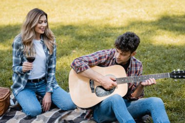 Kadın elinde kırmızı şarap kadehiyle akustik gitar çalan sevgilisine bakıyor. 