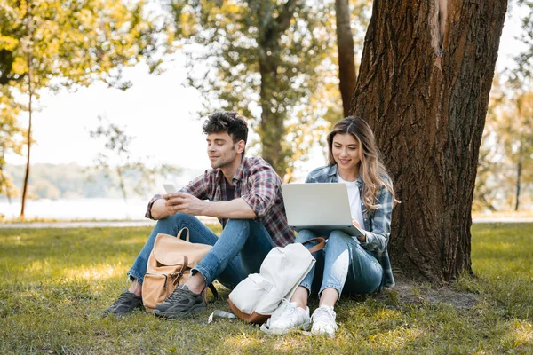 Casal Usando Laptop Smartphone Enquanto Sentado Sob Tronco Árvore — Fotografia de Stock