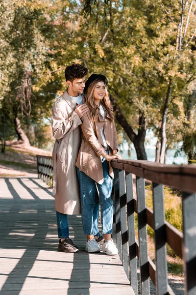 man touching shoulders of woman in hat and trench coat standing on wooden bridge in autumnal park