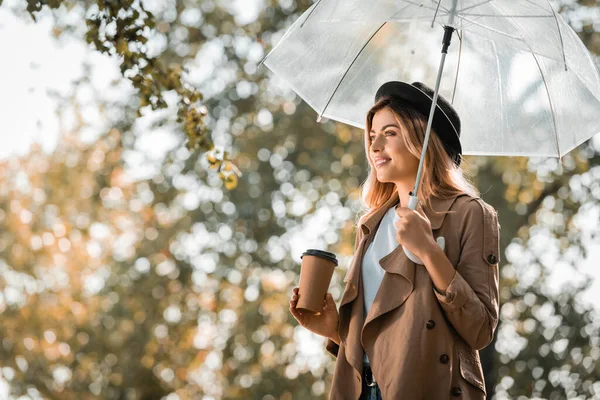 トレンチコートの女と傘の下に立つ帽子秋の公園で紙コップを持ち — ストック写真