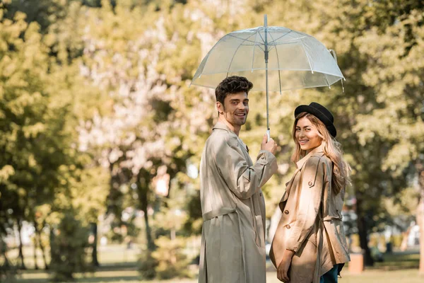 blonde woman in hat and man standing under umbrella in autumnal park