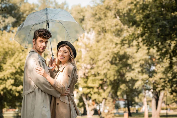帽子のブロンド女性と傘の下に抱きかかえているトレンチコートの男秋の公園でカメラを見て — ストック写真