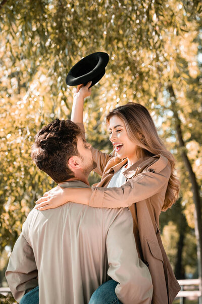 man holding in arms excited woman with hat in autumnal park