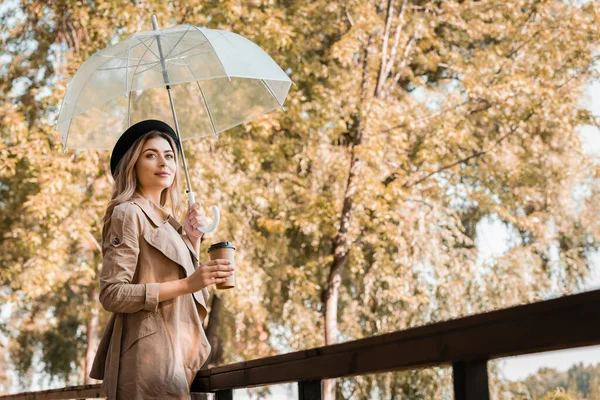 秋の公園に行くために傘とコーヒーと使い捨てカップを持って帽子の女性 — ストック写真