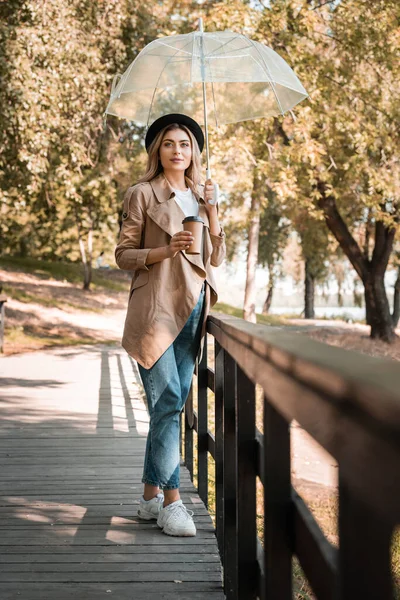 秋の公園の橋の上に立っている間に行くためにコーヒーと傘と使い捨てカップを保持帽子の女性の選択的な焦点 — ストック写真