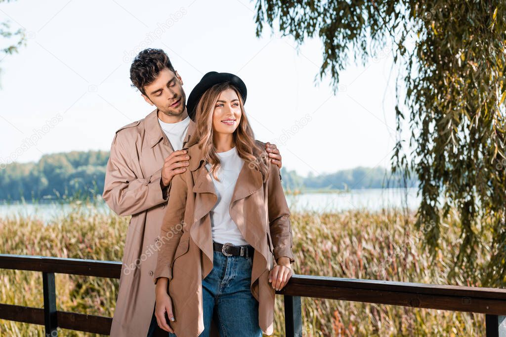 man touching shoulders of woman in hat and trench coat in autumnal park