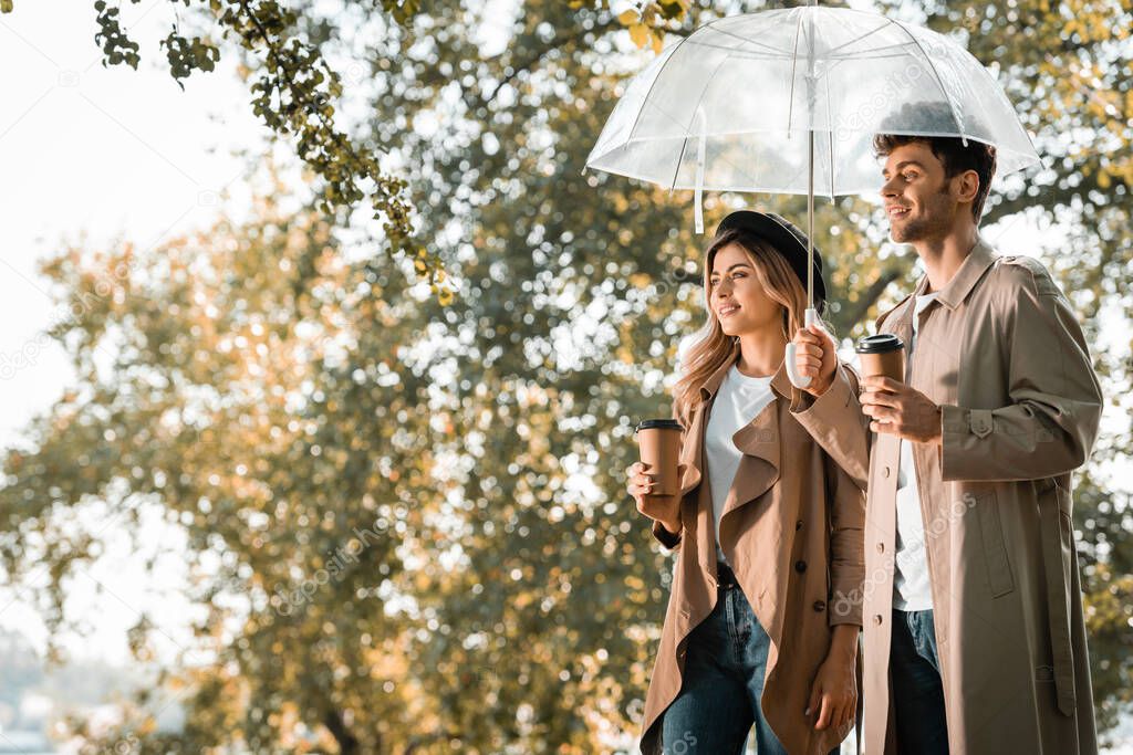 couple in trench coats standing under umbrella and holding paper cups with coffee to go