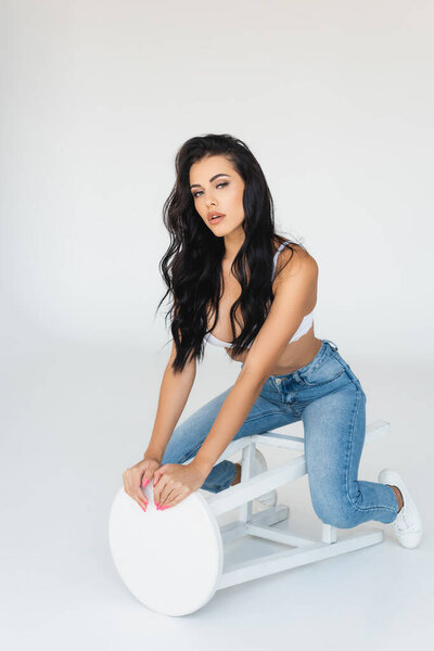 сексуальная брюнетка в джинсах и лифчике сидящая и трогательная табуретка на сером