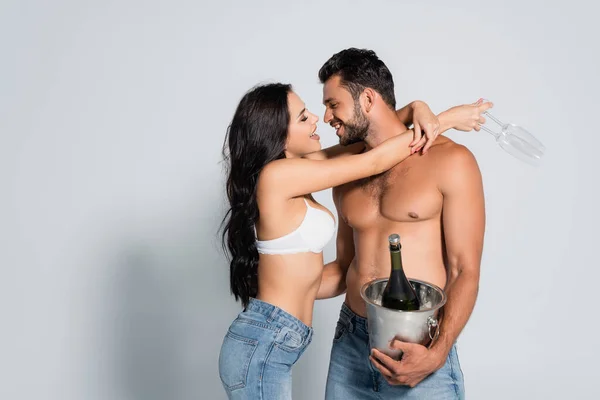 seductive woman in bra holding glasses and hugging shirtless man with ice bucket and bottle of champagne on grey