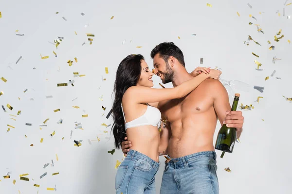 brunette woman in bra hugging muscular man with bottle of champagne on grey with confetti