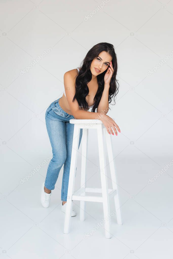 Sexy woman in bra and jeans leaning on stool and looking at camera on grey