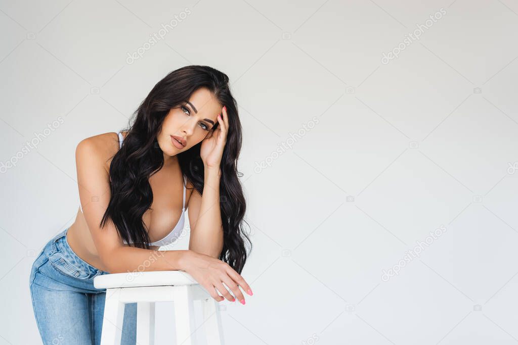 sexy young woman in bra and jeans leaning on chair and looking at camera on grey