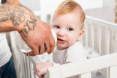 selective focus of tattooed man feeding infant child standing in cot 