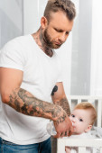 mladý tetovaný muž krmení dítě stojící v postýlce